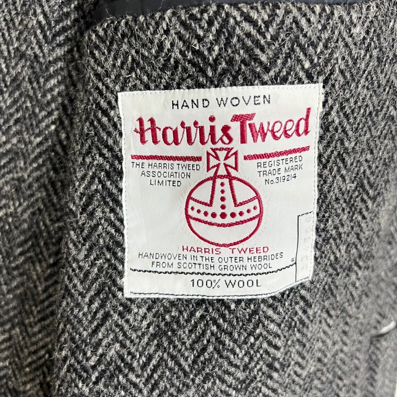 Puede incluir: Primer plano de una tela de tweed gris y negro con dise&ntilde;o de espiga. Una etiqueta blanca con texto rojo dice "Harris Tweed", con detalles sobre su origen y que est&aacute; tejida a mano con lana escocesa.