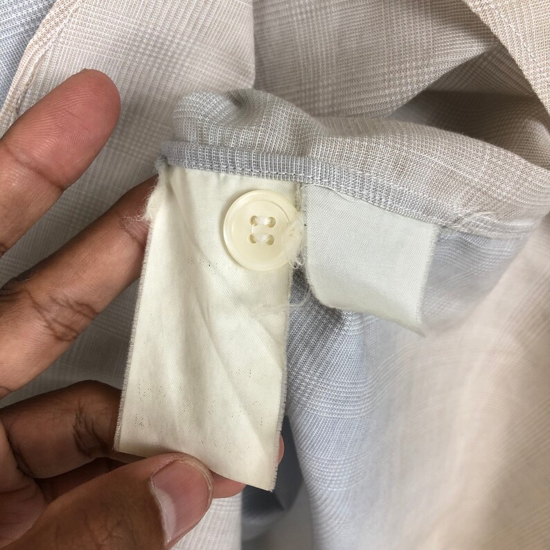 Op de afbeelding: Een close-up van een wit stoffen label met een erop gestikte knoop. Het label is bevestigd aan een lichtgrijs en wit geruit overhemd.