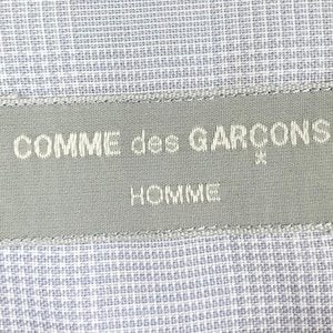 Op de afbeelding: Een grijs stoffen label met de tekst "COMME des GARCONS* HOMME" in wit.