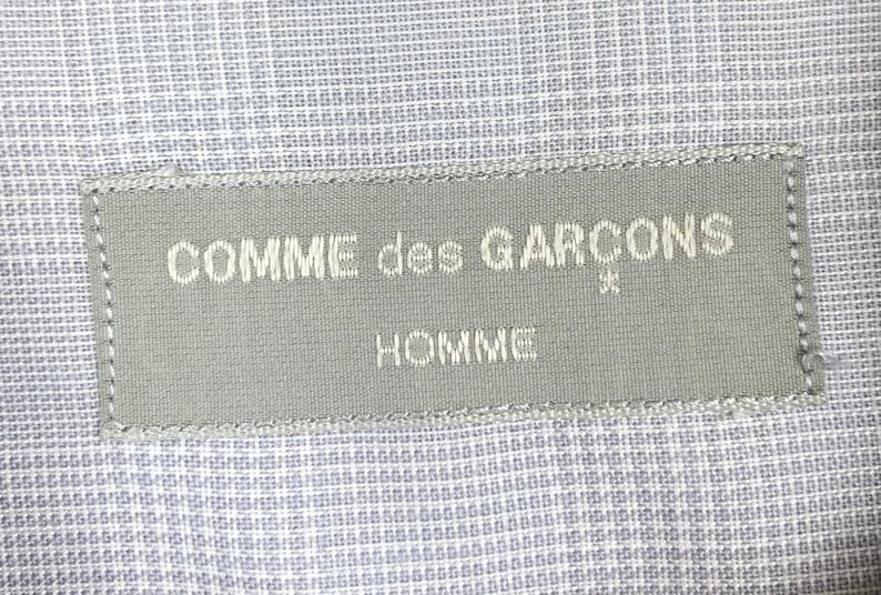 Op de afbeelding: Een grijs stoffen label met de tekst "COMME des GARCONS* HOMME" in wit.
