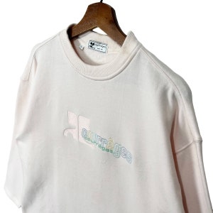 May include: Pale pink sweatshirt with the Courr&egrave;ges logo embroidered in pastel colours. The sweatshirt is hanging on a wooden hanger. The label reads "Courr&egrave;ges Paris".