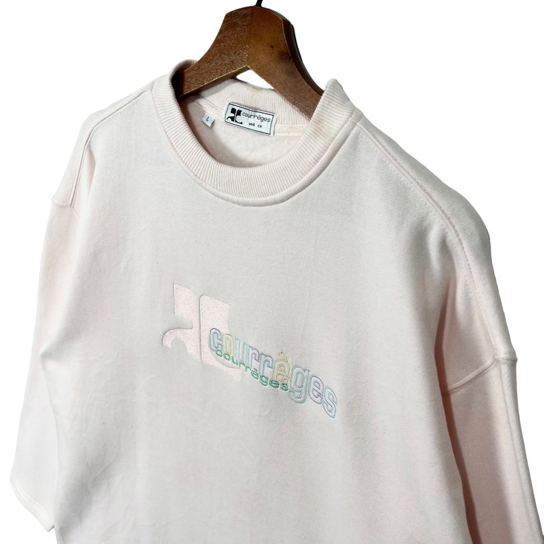 May include: Pale pink sweatshirt with the Courr&egrave;ges logo embroidered in pastel colours. The sweatshirt is hanging on a wooden hanger. The label reads "Courr&egrave;ges Paris".