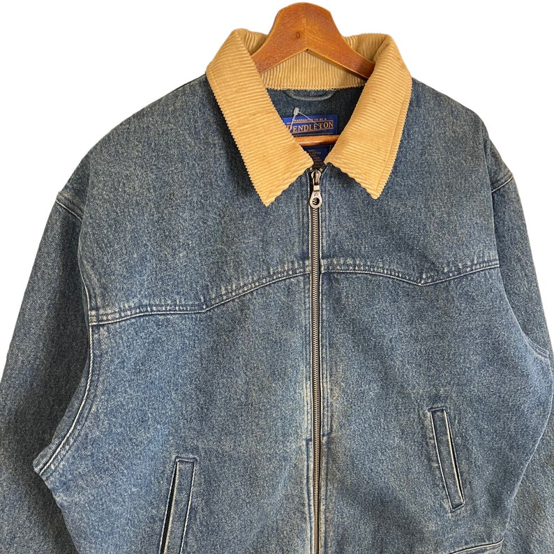 Pode incluir: Casaco de ganga azul com gola de veludo cotel&ecirc; bege. O casaco tem fecho de correr completo, dois bolsos frontais e uma etiqueta da marca "Pendleton". Est&aacute; pendurado num cabide de madeira.