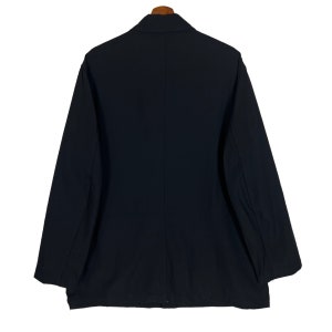 Op de afbeelding: Een zwarte blazer hangt aan een houten kleerhanger. De jas heeft lange mouwen en een klassieke kraag. De achterkant van de blazer toont een strak, minimalistisch ontwerp, geschikt voor diverse gelegenheden. De stof lijkt een effen, gladde kleur te hebben.