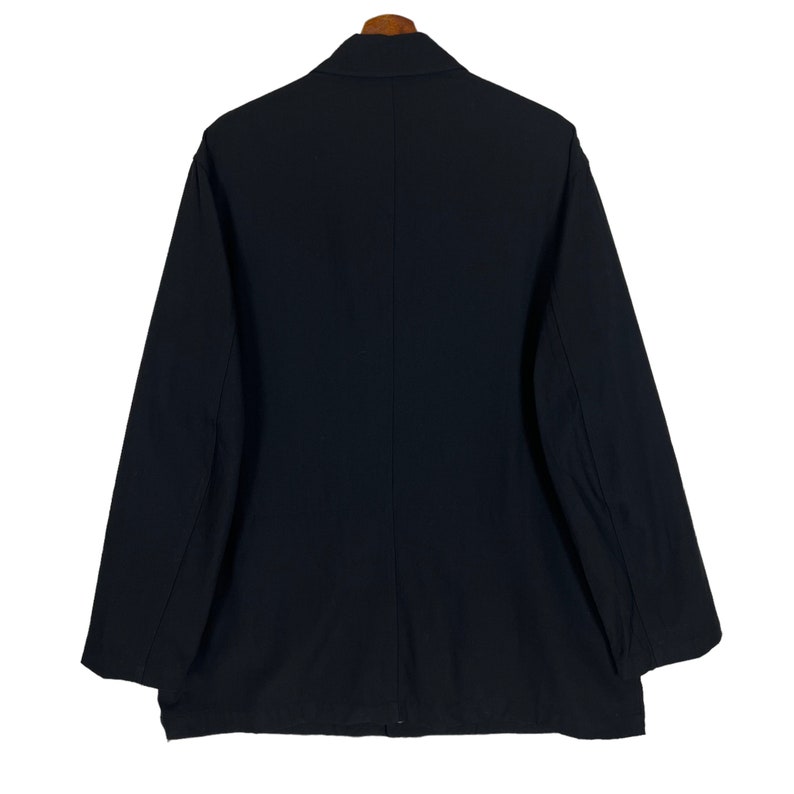 Op de afbeelding: Een zwarte blazer hangt aan een houten kleerhanger. De jas heeft lange mouwen en een klassieke kraag. De achterkant van de blazer toont een strak, minimalistisch ontwerp, geschikt voor diverse gelegenheden. De stof lijkt een effen, gladde kleur te hebben.