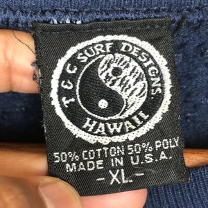 Pu&ograve; includere: Un'etichetta in bianco e nero con il testo "I.C. Surf Designs Hawaii" in un cerchio con un simbolo yin yang al suo interno. L'etichetta dice anche "50% Cotone 50% Poli Made in U.S.A. - XL -"