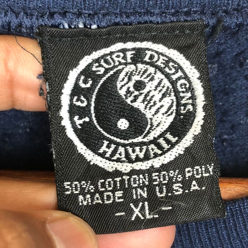 Pu&ograve; includere: Un'etichetta in bianco e nero con il testo "I.C. Surf Designs Hawaii" in un cerchio con un simbolo yin yang al suo interno. L'etichetta dice anche "50% Cotone 50% Poli Made in U.S.A. - XL -"