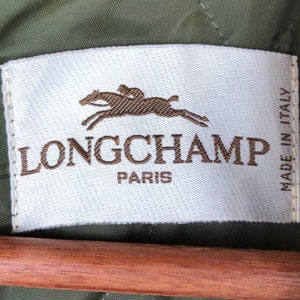 Pu&ograve; includere: Un'etichetta bianca con il marchio "LONGCHAMP PARIS" in lettere marroni. Un logo marrone di un cavallo e un cavaliere &egrave; sopra il marchio. L'etichetta &egrave; su uno sfondo di tessuto verde, con "MADE IN ITALY" stampato verticalmente.