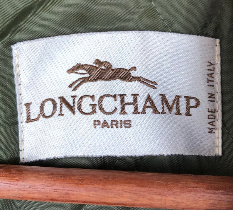 Pu&ograve; includere: Un'etichetta bianca con il marchio "LONGCHAMP PARIS" in lettere marroni. Un logo marrone di un cavallo e un cavaliere &egrave; sopra il marchio. L'etichetta &egrave; su uno sfondo di tessuto verde, con "MADE IN ITALY" stampato verticalmente.