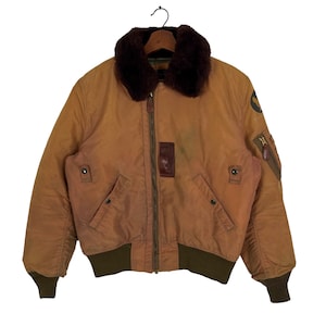 Puede incluir: Una chaqueta bomber de cuero marrón con cuello de piel de oveja y cierre de cremallera. La chaqueta tiene un parche en la manga izquierda con el texto "U.S. AIR FORCE".