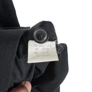 May include: A black fabric label with white text that reads 'YN-D51-113' and 'Wool 100%'. The label also has the text 'Lining' and 'Cupro' in Japanese. The label has the text 'Dry Clean Only' in Japanese.