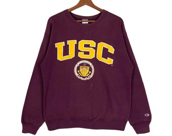 May include: Burgundy sweatshirt with a large yellow "USC" logo and a smaller crest featuring the University of Southern California's seal. The sweatshirt has a Champion logo on the left sleeve.