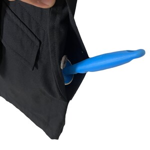 Op de afbeelding: Close-up van een zwarte kledingzak met een blauwe tool erin. De tool heeft een blauwe handgreep en een witte cirkelvormige basis. Er is ook een zilveren munt zichtbaar in de zak.