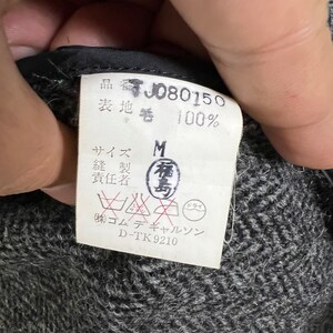 Puede incluir: Primer plano de una etiqueta de ropa con texto japon&eacute;s y el n&uacute;mero "J080150". La etiqueta indica "100%" de material y una talla "M". La etiqueta tambi&eacute;n incluye instrucciones de lavado y el nombre de la marca "コムデギャルソン".