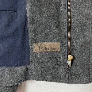 May include: A gray wool jacket with a zipper closure and a black fabric patch on the left side. The patch has the text "Y's for men" printed on it.