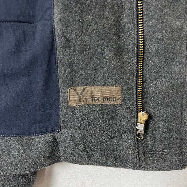 May include: A gray wool jacket with a zipper closure and a black fabric patch on the left side. The patch has the text "Y's for men" printed on it.