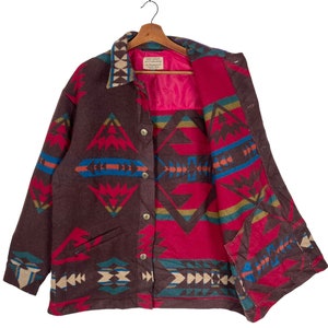 May include: A brown, button-down jacket with a Southwestern geometric pattern in red, blue, and beige. The jacket has a pink lining and a collar with a patterned trim. The jacket is hanging on a wooden hanger.