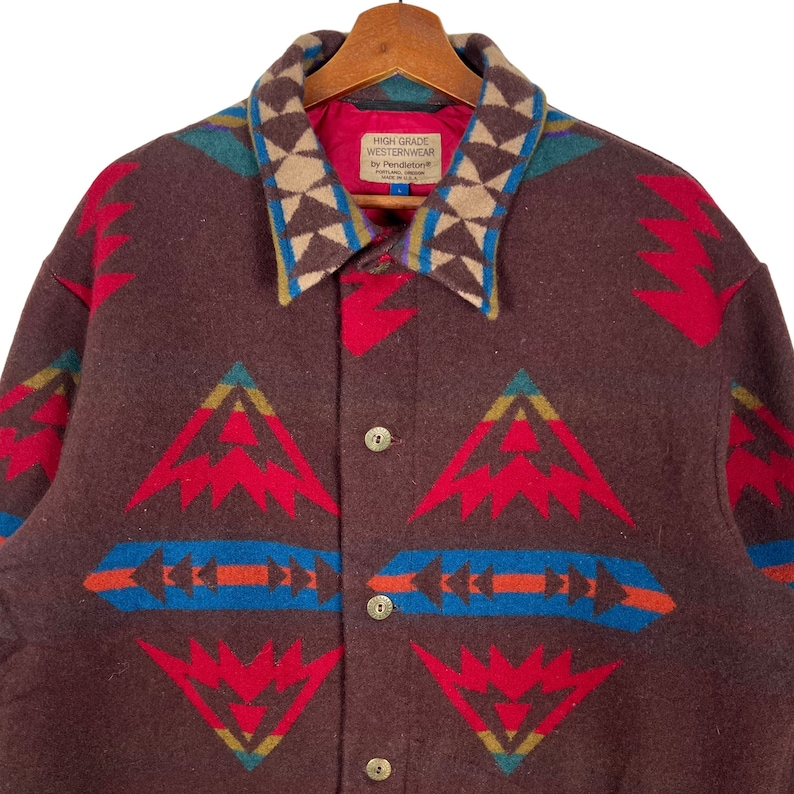 May include: A brown wool jacket with a colorful geometric pattern in red, blue, and tan. The jacket has a collared neckline and button closures. The label reads "HIGH GRADE WESTERNWEAR by Pendleton&reg;".