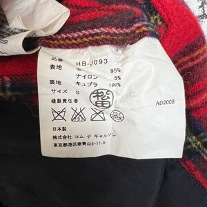 May include: A white tag with black text and symbols. The tag is attached to a red and black plaid fabric. The tag lists the fabric composition as 95% nylon and 5% cupro. The tag also includes the size, S, and the brand name, "Commes des Garcons".