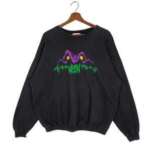 May include: Black crew neck sweatshirt with a purple and yellow graphic of a creature's eyes and mouth. The word "WEEN" is printed in green below the graphic, with a green heartbeat line. The sweatshirt is hanging on a wooden hanger.