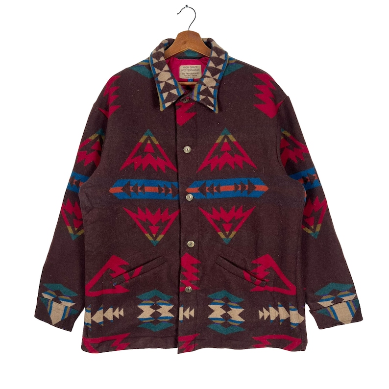 May include: A brown button-up jacket with a geometric pattern in red, blue, and yellow. The jacket has a collared neckline and two front pockets. The collar and cuffs have a patterned trim. The jacket is hanging on a wooden hanger.