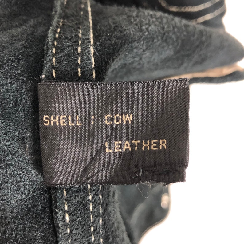 Puede incluir: Primer plano de una etiqueta de cuero negro con costuras blancas. La etiqueta dice "SHELL: COW LEATHER".