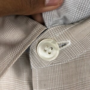 Op de afbeelding: Een close-up van een witte knoop met vier gaten, genaaid op een lichtbruine geruite stof.