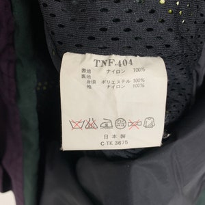 May include: A white tag with black text detailing the fabric composition of a black jacket. The tag reads "TNF.404", "表地 ナイロン 100%", "裏地 身頃 ポリエステル 100%", "袖 ナイロン 100%", "日本製", "C-TK 3675".