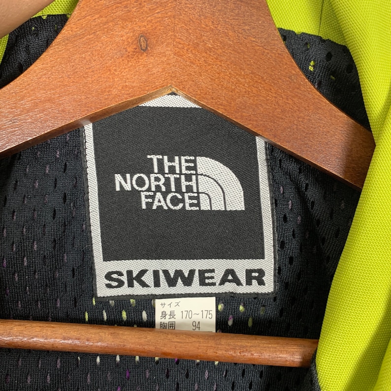 May include: Black and white The North Face skiwear label with white text and a white logo. The label has the text "Skiwear" in a large white font and the text "The North Face" in a smaller white font. The label also has a size tag with the text "Size: 170-175" and "94".