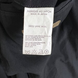 Op de afbeelding: Een wit kledinglabel met zwarte tekst, waaronder "FABRIQUE AU JAPON MADE IN JAPAN" en "100% LAINE WOOL". Het label bevat ook onderhoudsinstructies en symbolen voor chemisch reinigen, wassen en strijken. Het label is bevestigd aan een donker kledingstuk.