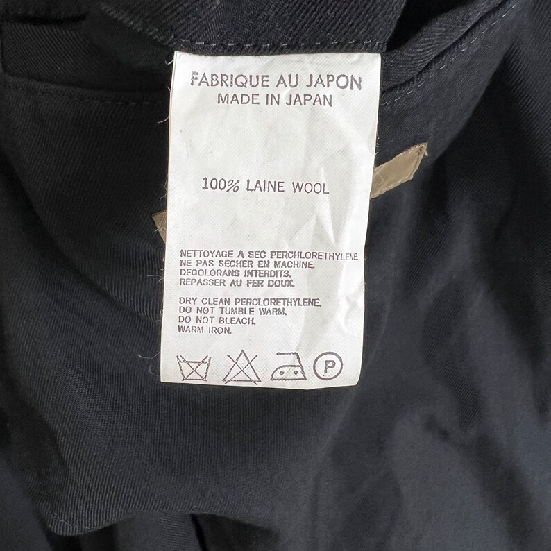 Op de afbeelding: Een wit kledinglabel met zwarte tekst, waaronder "FABRIQUE AU JAPON MADE IN JAPAN" en "100% LAINE WOOL". Het label bevat ook onderhoudsinstructies en symbolen voor chemisch reinigen, wassen en strijken. Het label is bevestigd aan een donker kledingstuk.