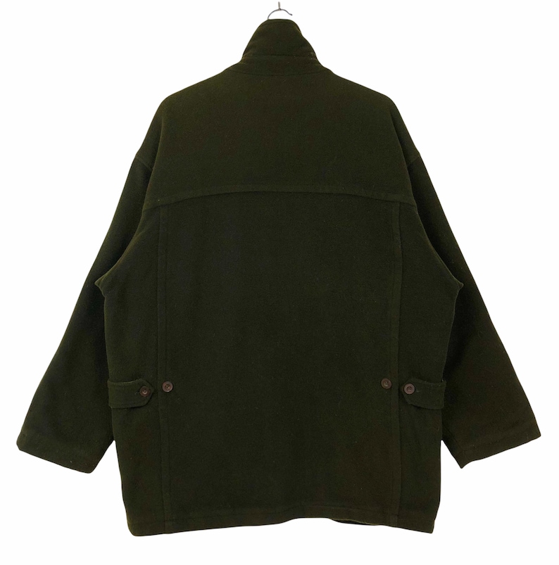 Pu&ograve; includere: Cappotto in lana verde oliva scuro con collo alto e due tasche frontali. Il cappotto ha una chiusura con bottoni e un design classico e oversize. Il cappotto &egrave; appeso a una gruccia di metallo su sfondo bianco.