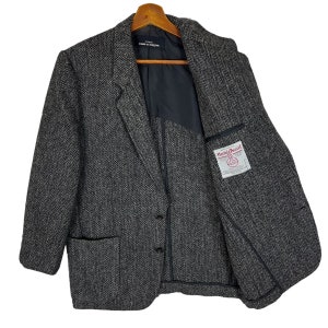 Puede incluir: Un blazer de tweed gris con dise&ntilde;o de espiga, cierre de dos botones y solapa con muescas. La chaqueta tiene dos bolsillos delanteros y una etiqueta dentro del bolsillo izquierdo del pecho. El blazer est&aacute; colgado de una percha de madera.