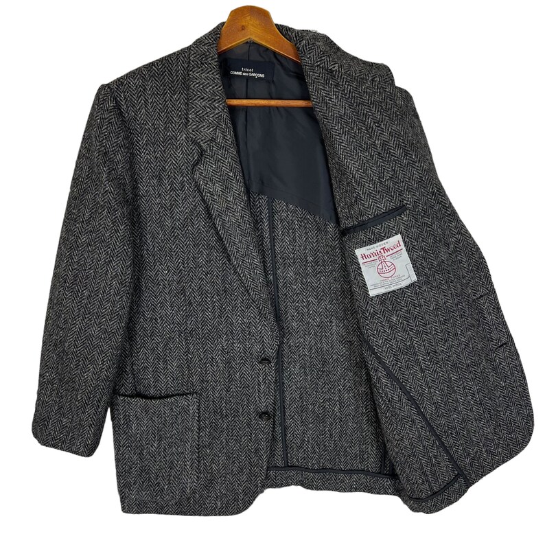 Puede incluir: Un blazer de tweed gris con dise&ntilde;o de espiga, cierre de dos botones y solapa con muescas. La chaqueta tiene dos bolsillos delanteros y una etiqueta dentro del bolsillo izquierdo del pecho. El blazer est&aacute; colgado de una percha de madera.