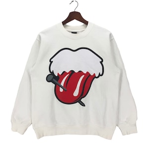 Puede incluir: Sudadera blanca con un gráfico de lengua y clavo rojo y blanco. El gráfico es una versión estilizada del logotipo de los Rolling Stones.