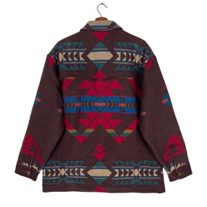 May include: A brown jacket with a colorful geometric pattern in red, blue, tan, and teal. The jacket is hanging on a wooden hanger. The design is symmetrical and covers the back of the jacket.