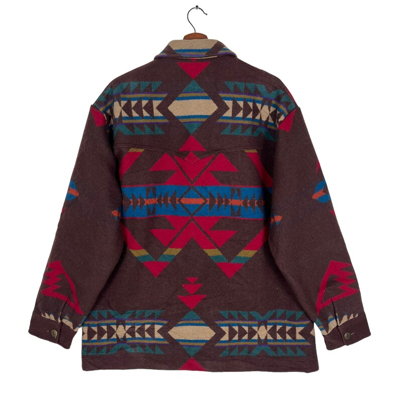 May include: A brown jacket with a colorful geometric pattern in red, blue, tan, and teal. The jacket is hanging on a wooden hanger. The design is symmetrical and covers the back of the jacket.