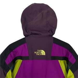 May include: Purple, black, and green color block jacket with a hood. The jacket has a black zipper and the logo "THE NORTH FACE" in yellow on the back.