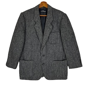 Puede incluir: Una blazer gris con dise&ntilde;o de espiga, dos bolsillos delanteros y cierre de dos botones. La blazer est&aacute; colgada en una percha de madera. La etiqueta dentro de la blazer dice "COMME des GAR&Ccedil;ONS".