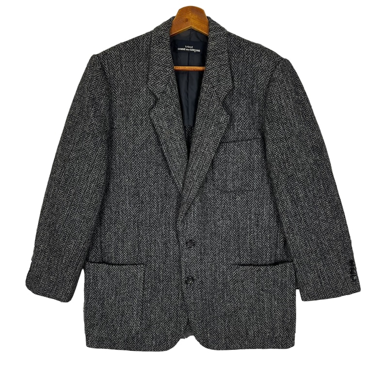 Puede incluir: Una blazer gris con dise&ntilde;o de espiga, dos bolsillos delanteros y cierre de dos botones. La blazer est&aacute; colgada en una percha de madera. La etiqueta dentro de la blazer dice "COMME des GAR&Ccedil;ONS".