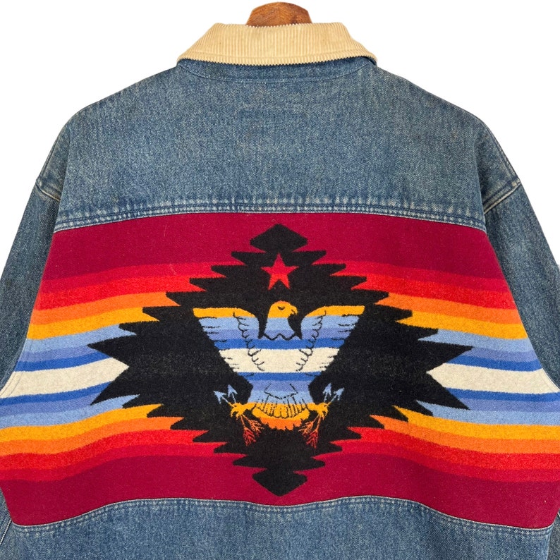 Pode incluir: Casaco de ganga azul com gola de veludo cotel&ecirc; creme. As costas apresentam um design colorido com uma &aacute;guia, uma estrela e padr&otilde;es geom&eacute;tricos em vermelho, laranja, amarelo, azul e preto.