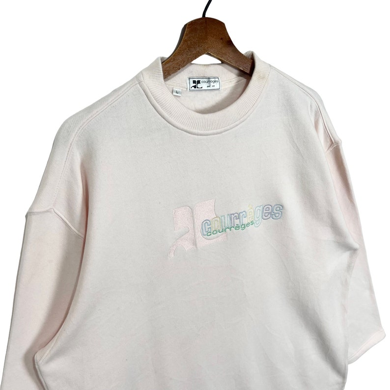 May include: Pale pink sweatshirt with the embroidered word "courr&egrave;ges" in a pastel colour scheme. The sweatshirt has a crew neck and long sleeves. The brand logo is visible on the neckline.