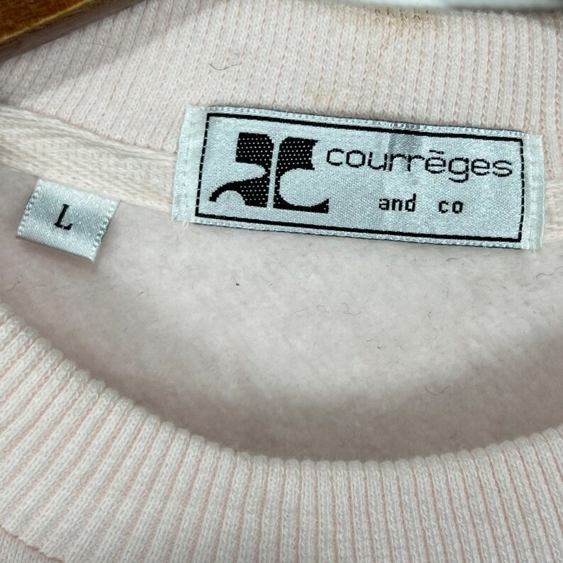 May include: A light pink sweatshirt with the brand label "courr&egrave;ges and co" in view. The label is rectangular with black text and a logo. A small tag with the letter "L" is also visible on the garment.