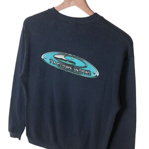 Pu&ograve; includere: Una felpa nera a maniche lunghe con una grafica blu e bianca sul retro che dice "TSC Surf Designs".