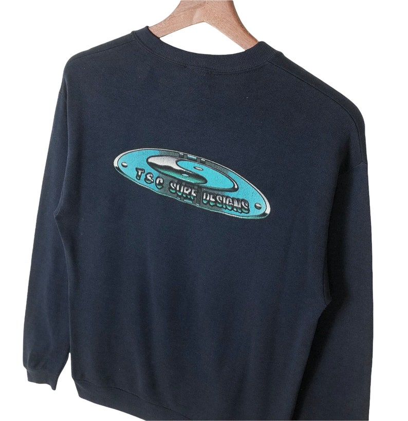 Pu&ograve; includere: Una felpa nera a maniche lunghe con una grafica blu e bianca sul retro che dice "TSC Surf Designs".