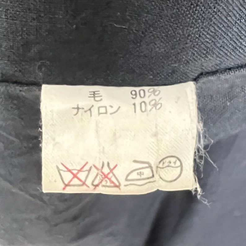 May include: A white fabric label with black text that reads "毛 90% ナイロン 10%". The label also has a red and black graphic of a washing tub with a red line through it, a red line through an iron, and a graphic of an iron with a dot inside a circle.