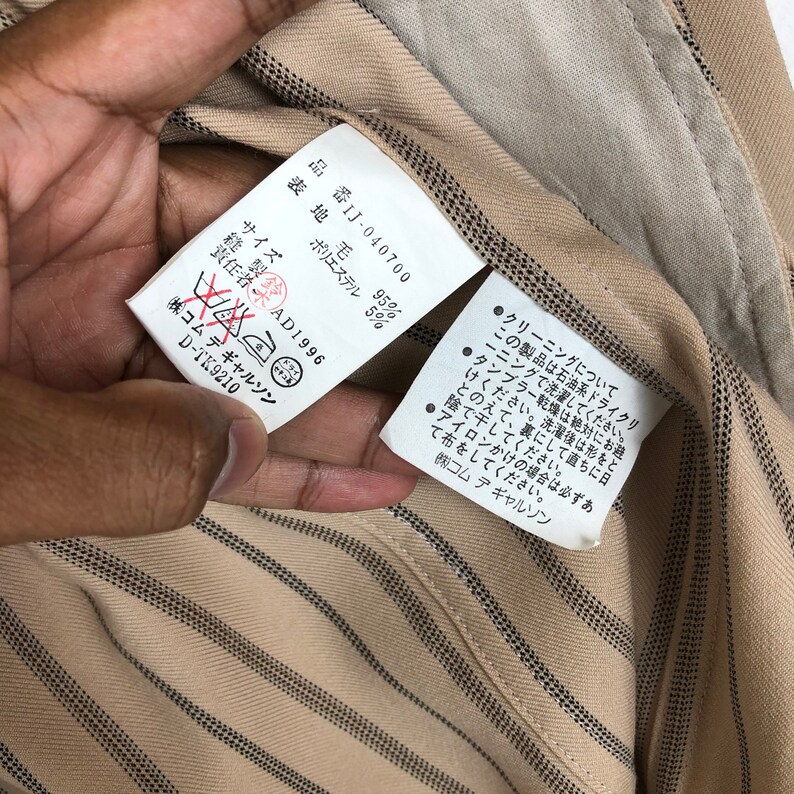 K&ouml;nnte beinhalten: Ein wei&szlig;es Etikett mit schwarzem Text, das die Stoffzusammensetzung und Pflegehinweise f&uuml;r ein gestreiftes Hemd angibt. Das Etikett lautet "表地毛 95% ポリエステル 5% 縫製鈴 責任者 AD1996 (株)コムデギャルソン D-TK9210" und enth&auml;lt ein Trockenreinigungssymbol.