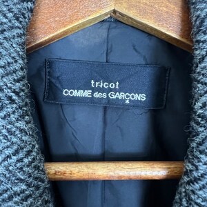 Puede incluir: Primer plano de una chaqueta gris y negra con estampado de espiga. El interior de la chaqueta presenta un forro negro y una etiqueta que dice "tricot COMME des GAR&Ccedil;ONS" en texto blanco. La chaqueta est&aacute; colgada en una percha de madera.