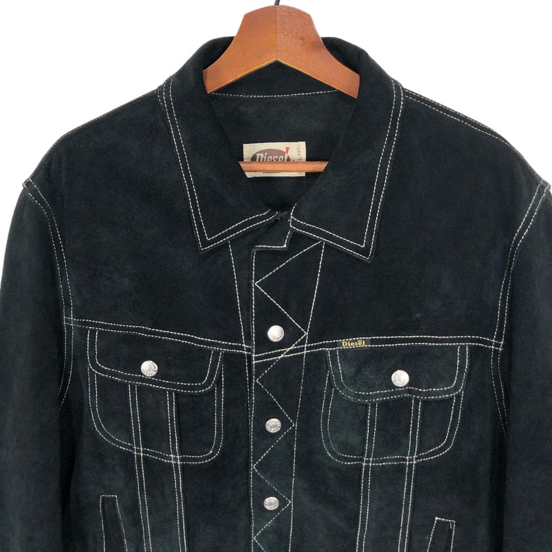 Puede incluir: Chaqueta de ante negro con costuras blancas y la marca Diesel. La chaqueta tiene cierre de botones y dos bolsillos de solapa.
