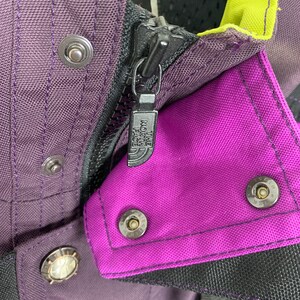 May include: Close-up of a purple and black jacket zipper with the The North Face logo. The zipper is metal and has a black pull tab. The jacket is made of a durable nylon fabric.
